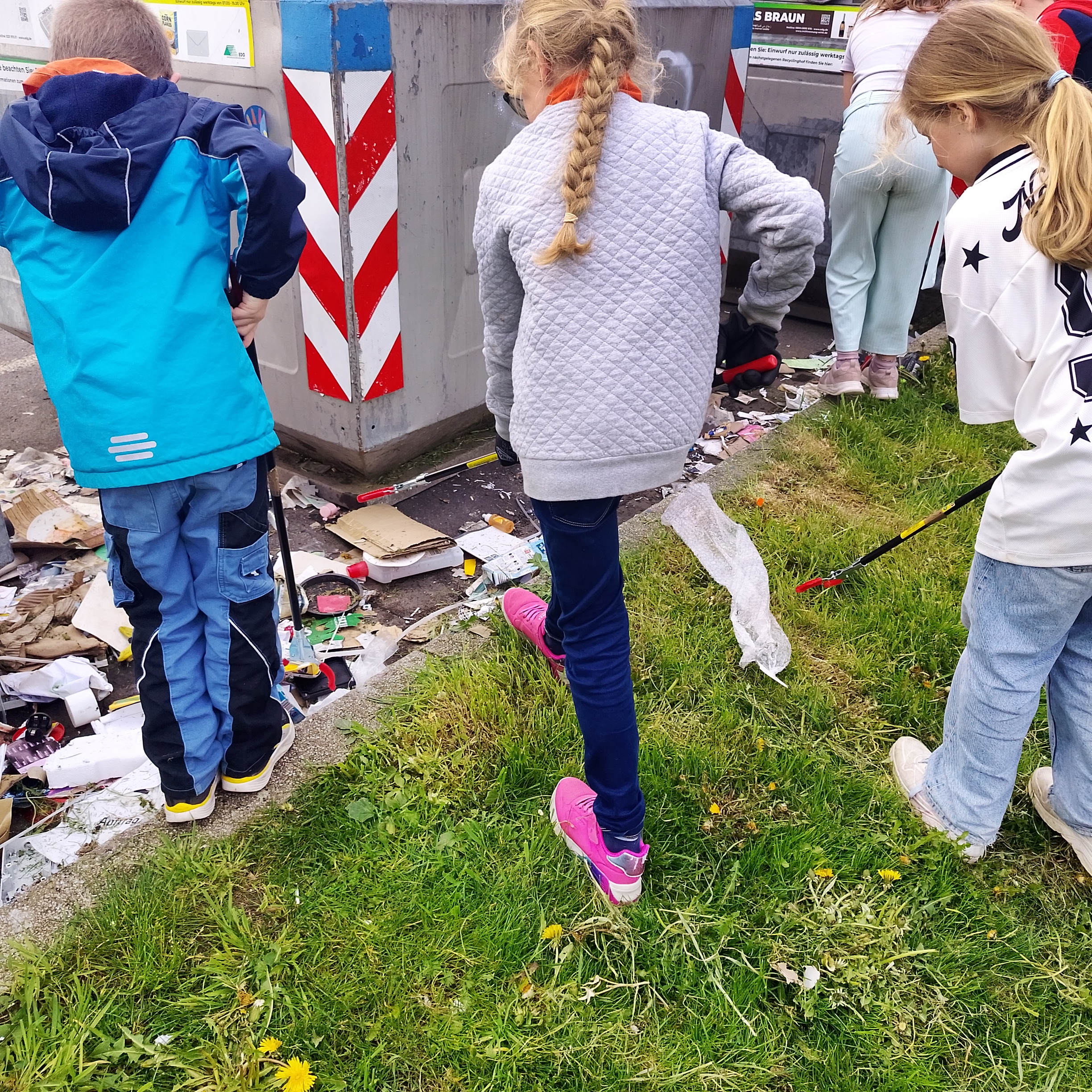 Mehrere Kinder stehen neben großen Müllcontainern und sammeln verstreuten Abfall vom Boden auf. Mit Greifzangen und Handschuhen heben sie Papier, Plastik und andere Reste auf, die rund um die Container liegen. Der Müll hat sich bis auf den angrenzenden Rasen ausgebreitet. Die Kinder arbeiten konzentriert zusammen und kümmern sich darum, den Bereich wieder sauber zu machen.